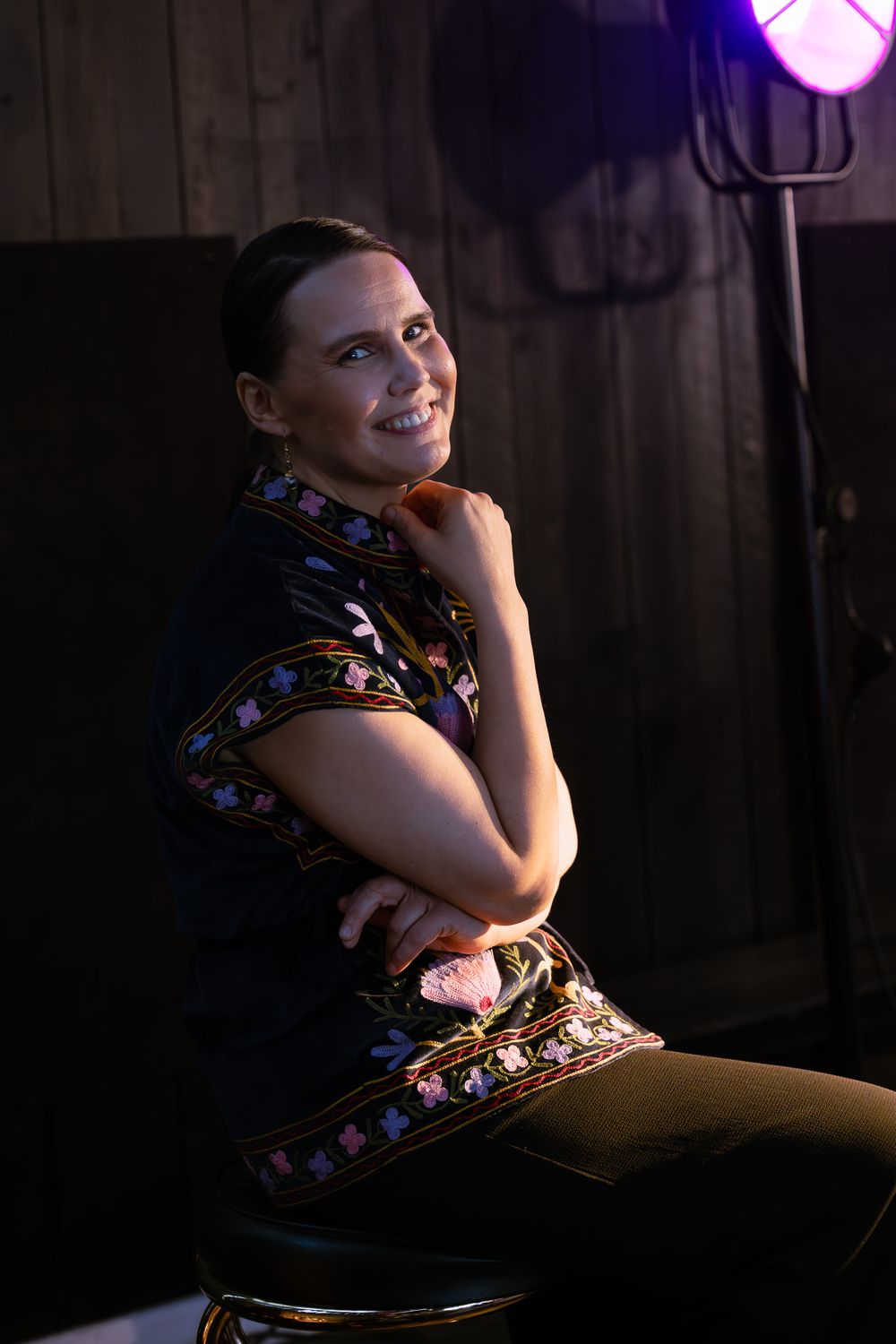 alt="A smiling woman in a dark embroidered shirt sits on a stool in a dimly lit studio with dramatic lighting"