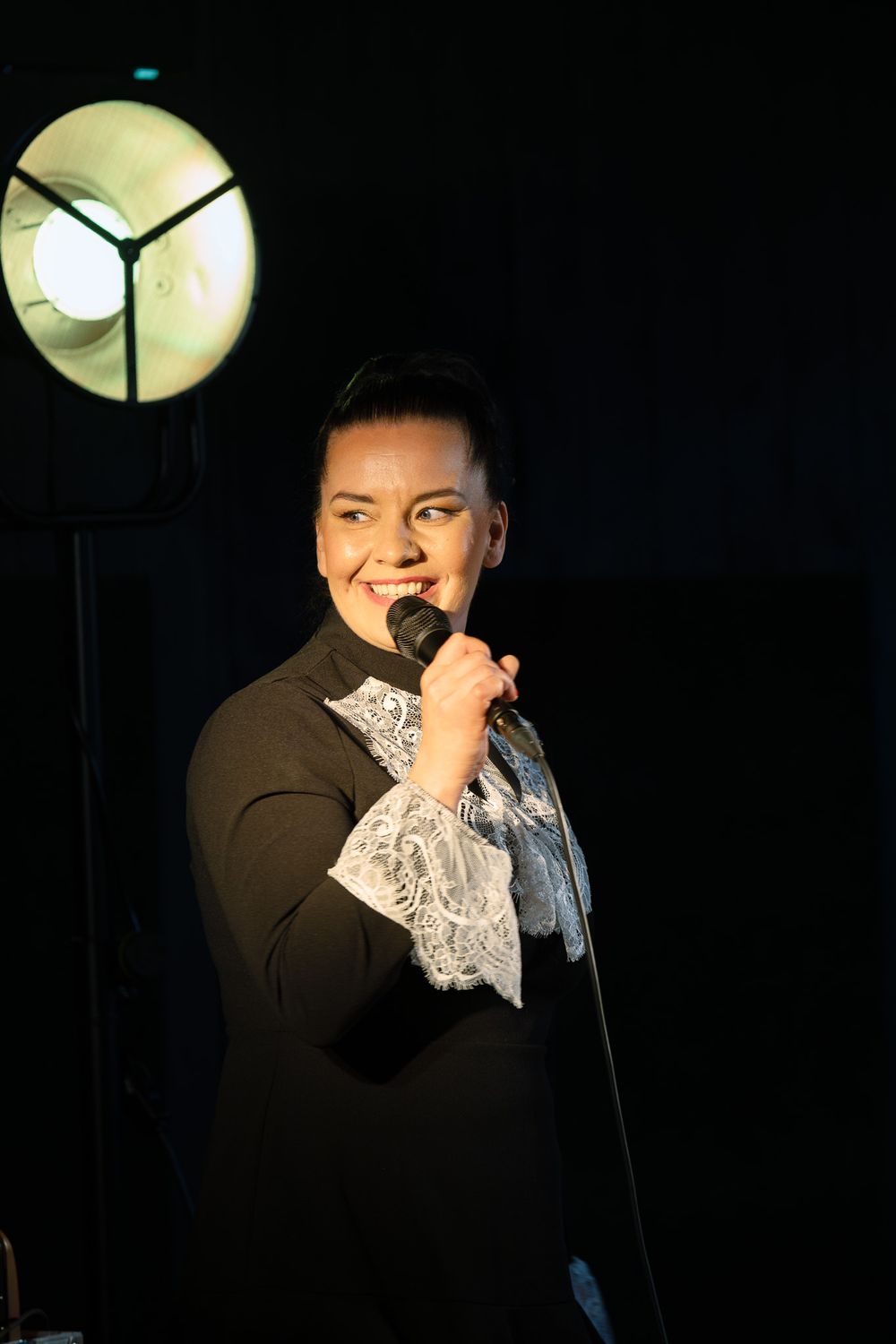 alt="Performer in black outfit with lace cuffs holds microphone on dark stage under spotlight, smiling confidently"