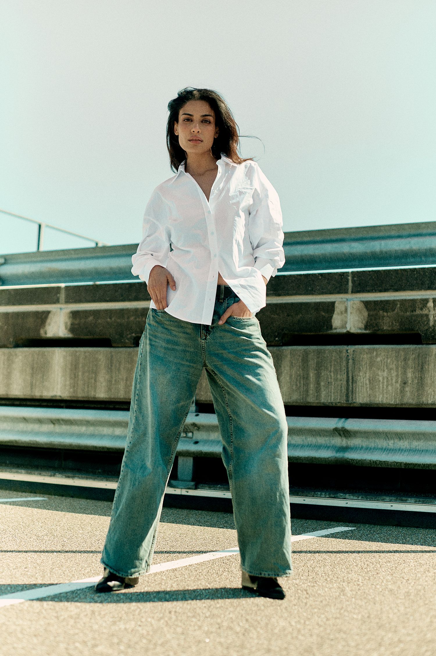 Woman in white oversized shirt and wide-leg jeans posing on rooftop parking lot in natural light.
