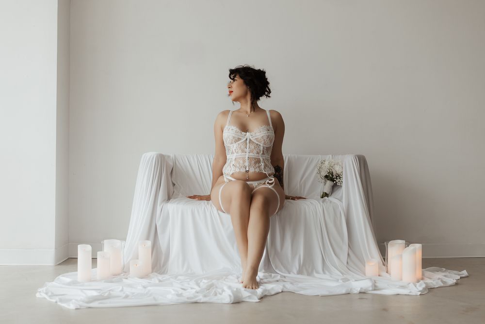 Woman in white lace lingerie corset sits on draped couch surrounded by pillar candles in bright minimalist studio.