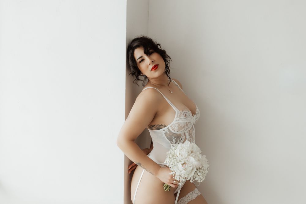 Brunette woman in white lace bodysuit lingerie holding white flower bouquet.