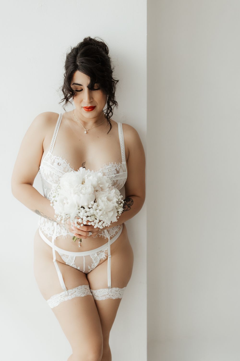 Curvy bride in white lace lingerie with garter belt holding a white peony bouquet against a light wall.