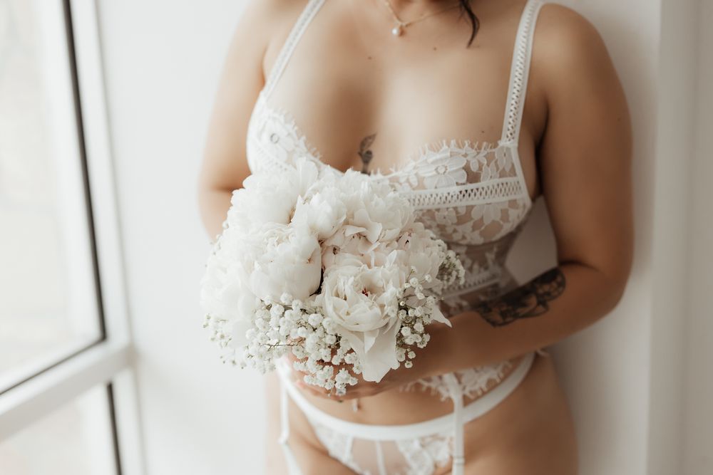 Close-up of a tattooed woman in white lace bridal lingerie holding a bouquet of white peonies and baby's breath.