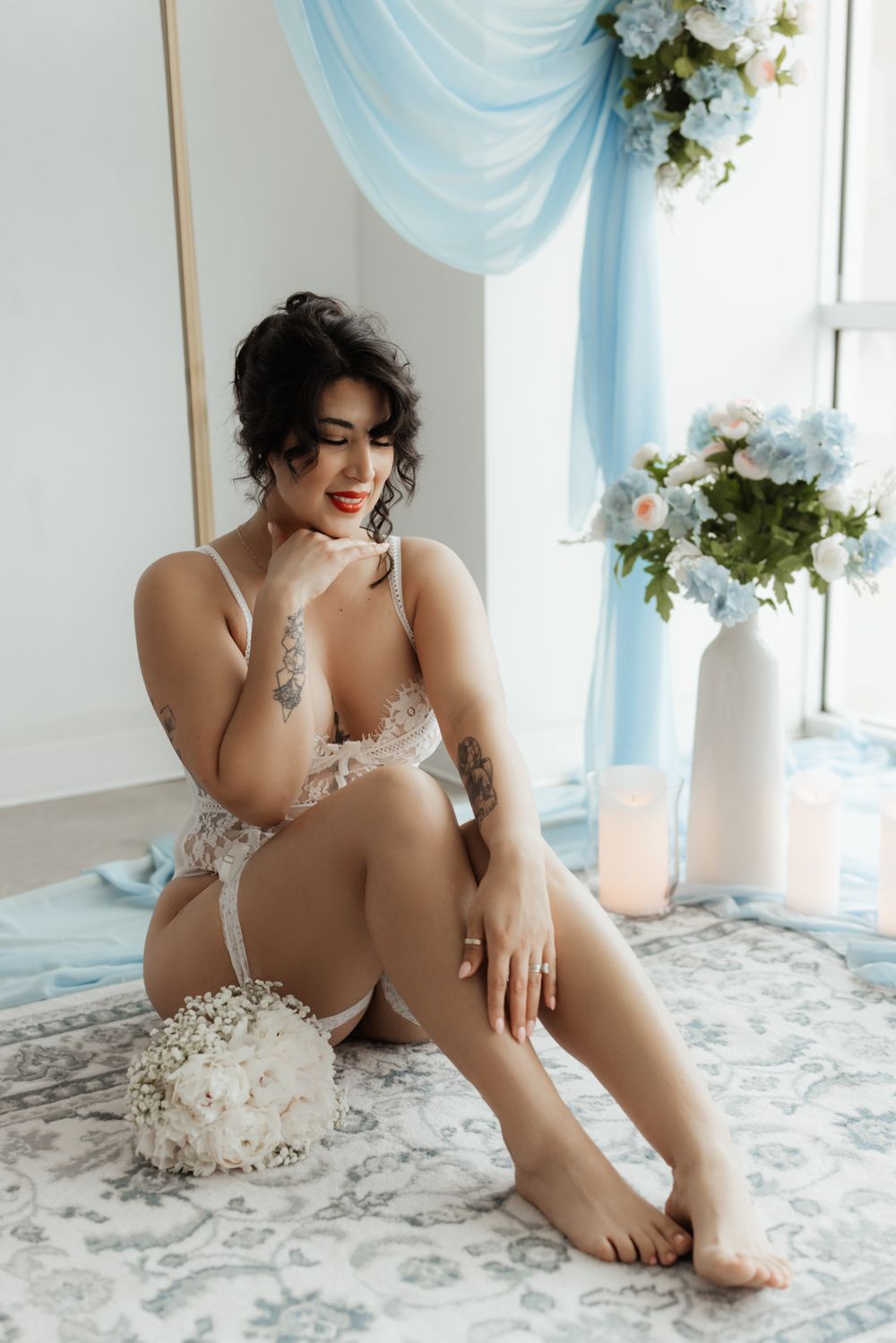 Woman in white lace lingerie sitting on floor with floral bouquet, blue drapes and flowers in bright studio setting.