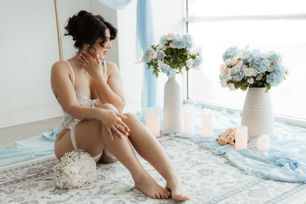 Woman in white lace lingerie sitting gracefully surrounded by blue and white floral arrangements indoors.