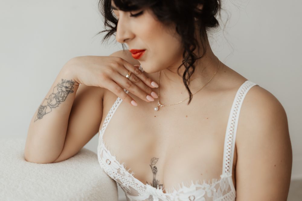Close-up of a dark-haired woman in white lace lingerie with red lips, tattoos, and delicate jewelry posing elegantly.