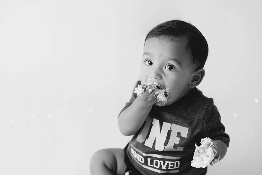 Black and white photo of a baby boy eating cake, wearing a shirt that reads ONE AND LOVED.