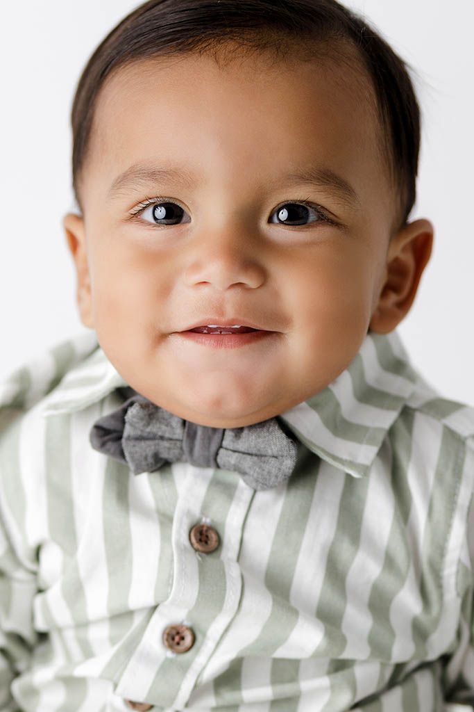 Adorable baby boy in striped green shirt and gray bow tie smiling at camera against white background.