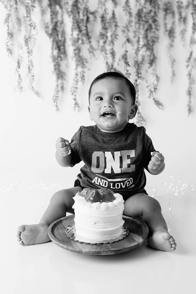 Black and white photo of a baby boy in a ONE AND LOVED onesie smiling at his first birthday cake smash Lake Elsinore photographer.