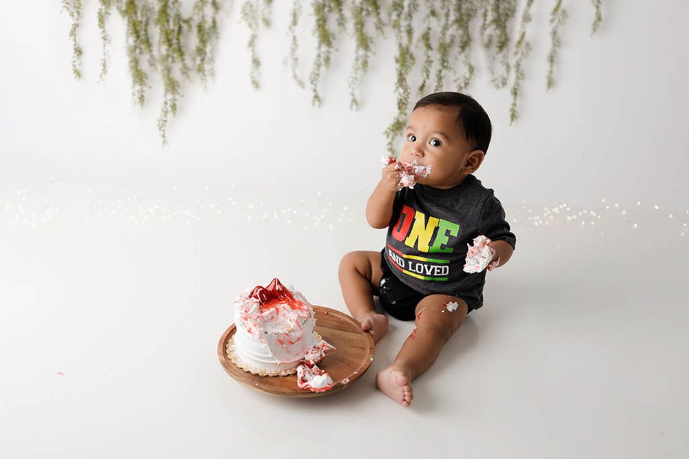 Baby in ONE onesie smashes first birthday cake on white backdrop with greenery.