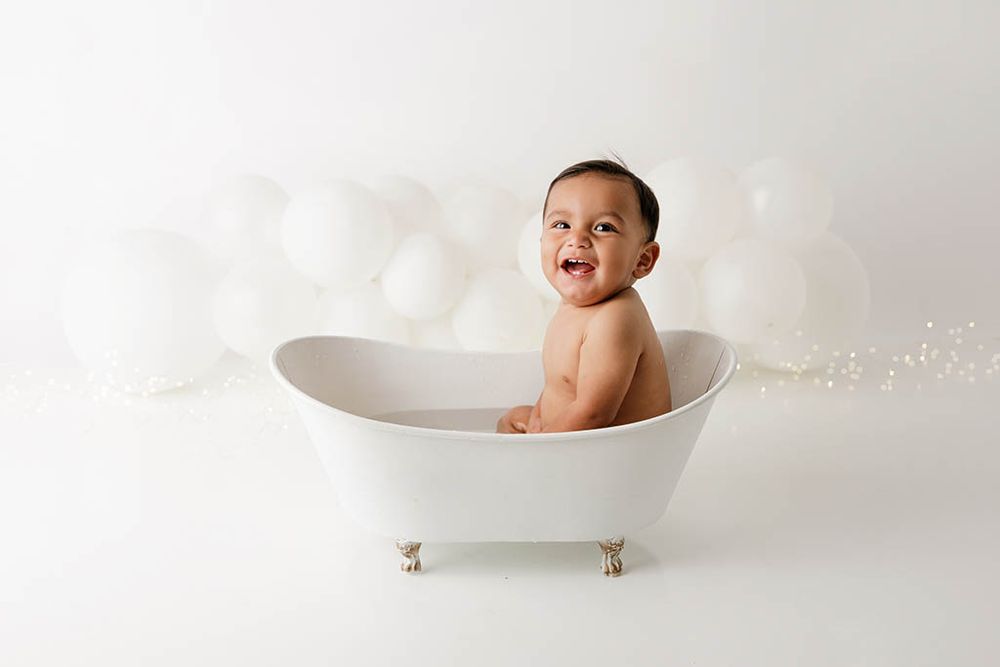 Happy baby sitting in a white clawfoot bathtub surrounded by white balloons in a bright studio setting.