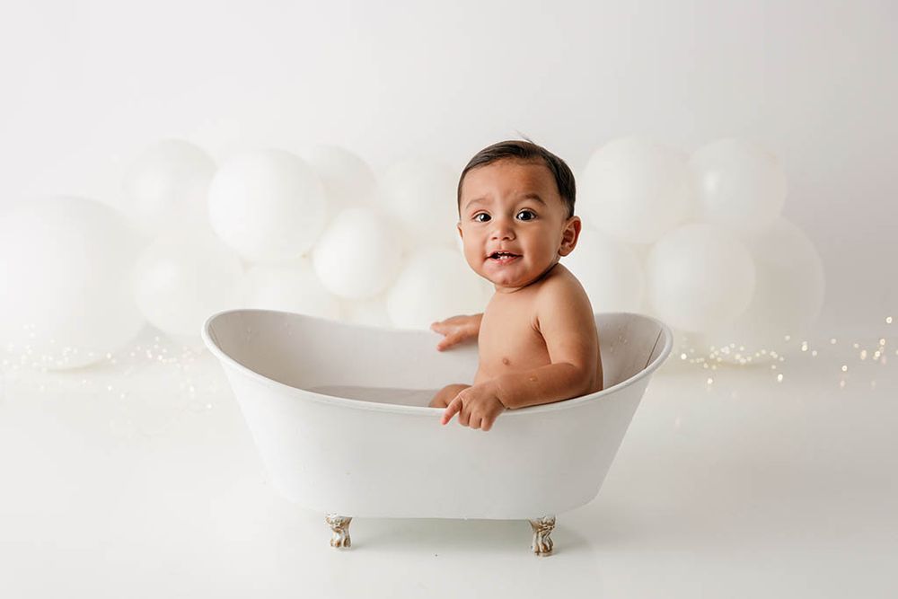 Smiling baby sitting in a bathtub surrounded by white balloons on a white background by professional Lake Elsinore 1st birthday photographer