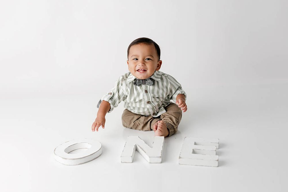 A smiling baby boy in a striped shirt sits with large white ONE letters for his first birthday near Lake Elsinore, California.