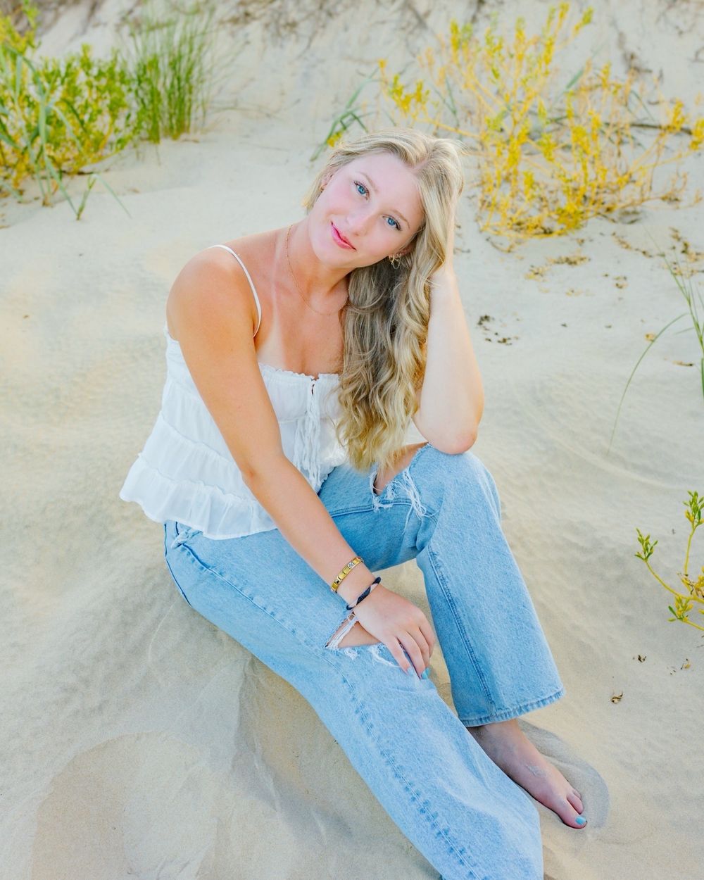 Blonde woman in white top and jeans sitting on sandy beach dunes with yellow wildflowers in background.