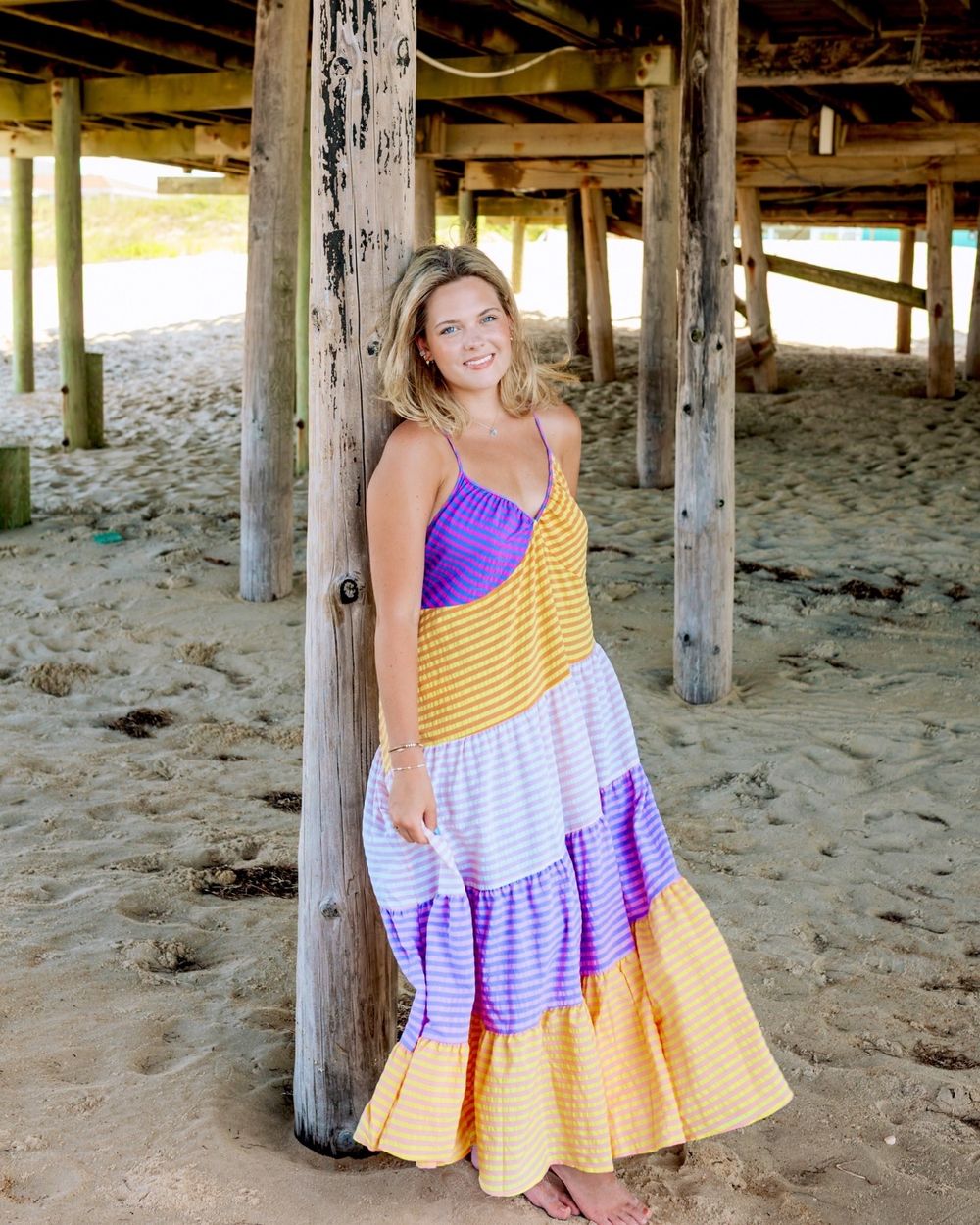 Woman in colorful striped maxi dress posing under beach pier on sandy shore.