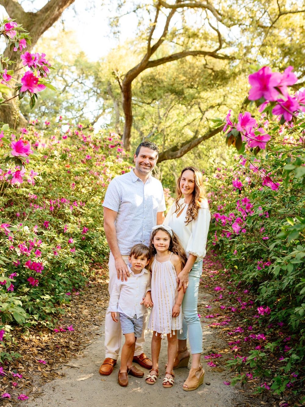 Family of four smiling on a garden path surrounded by vibrant pink azalea blooms and lush green trees.