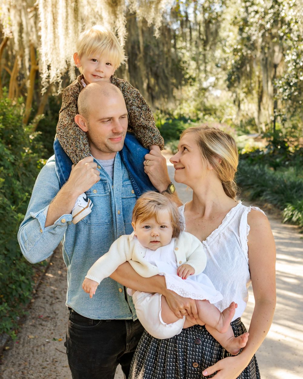 A happy family of four poses outdoors on a garden path surrounded by lush greenery and moss-draped trees.