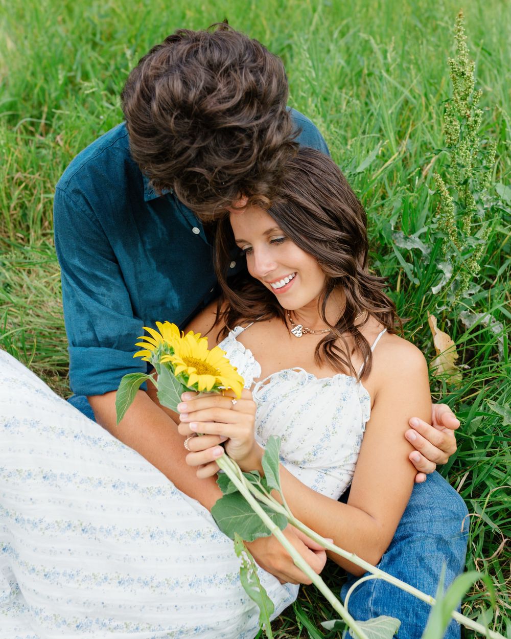 Couple sitting in green field, man in blue shirt embracing smiling woman holding yellow sunflower, romantic outdoor portrait.