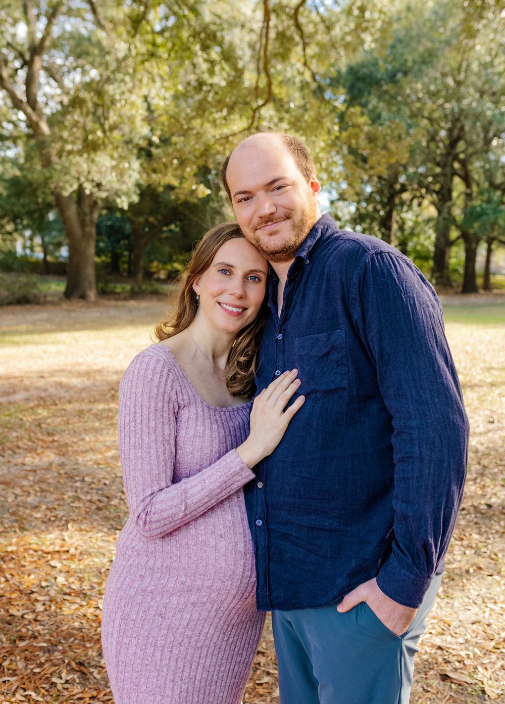 Pregnant woman in lavender dress poses with man in navy shirt outdoors among autumn trees.