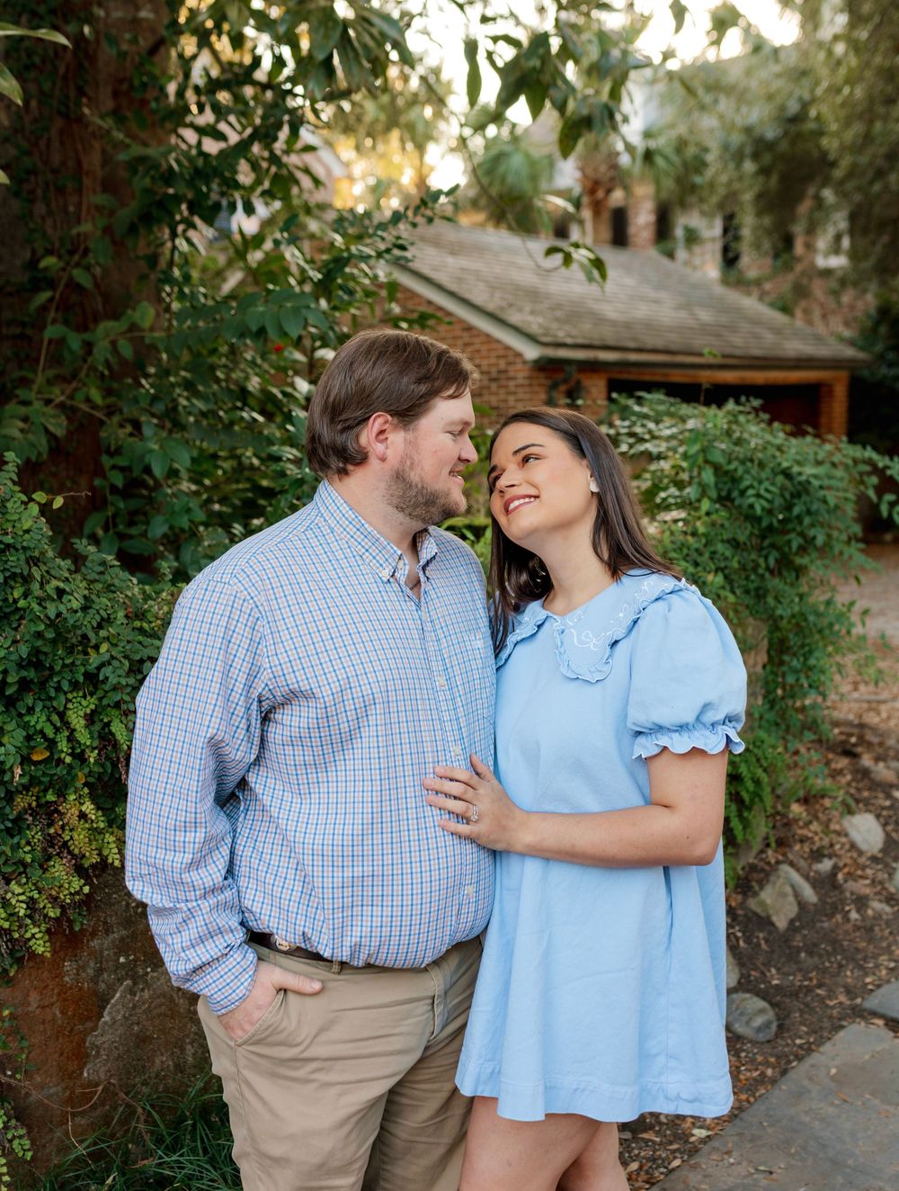 Couple in matching blue outfits share a romantic moment outdoors surrounded by lush greenery near a brick building.