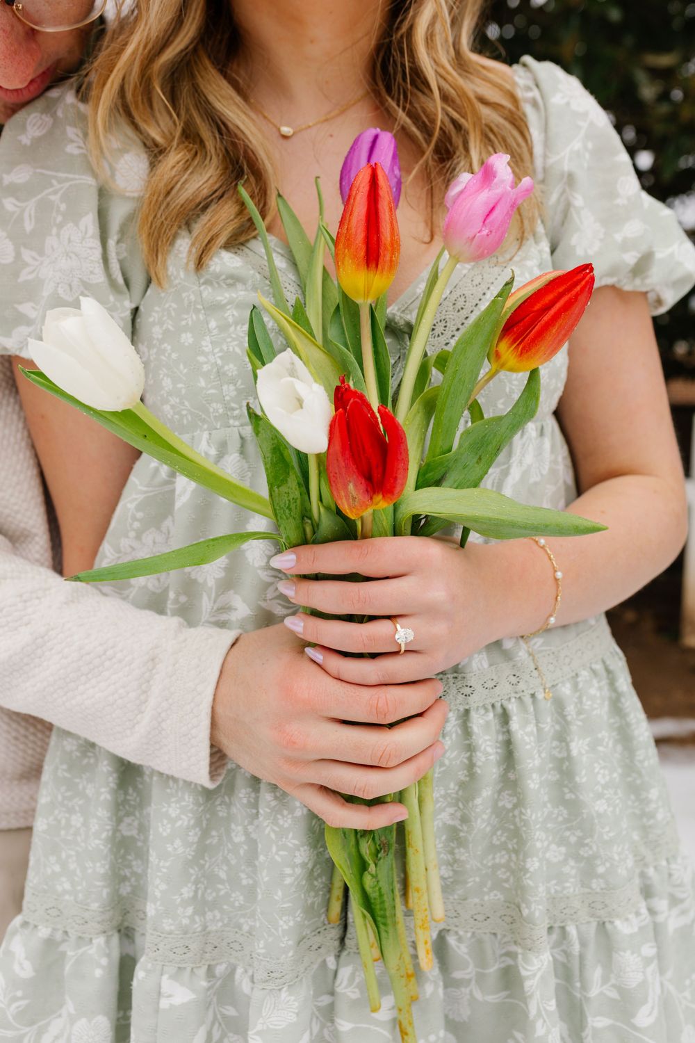 Woman in sage floral dress holding colorful tulip bouquet with diamond ring and gold bracelet.
