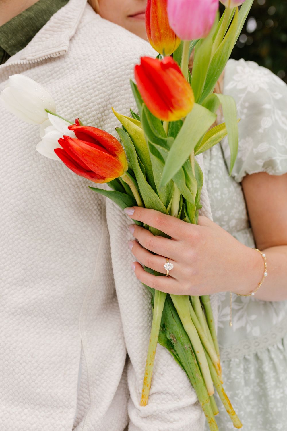 Close-up of hands holding colorful red, orange, and white tulips, with an engagement ring and gold bracelet visible.