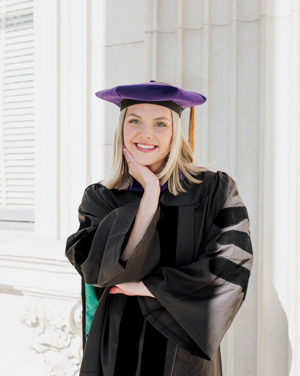 Smiling woman in black doctoral graduation gown and purple tam cap posing confidently outdoors.