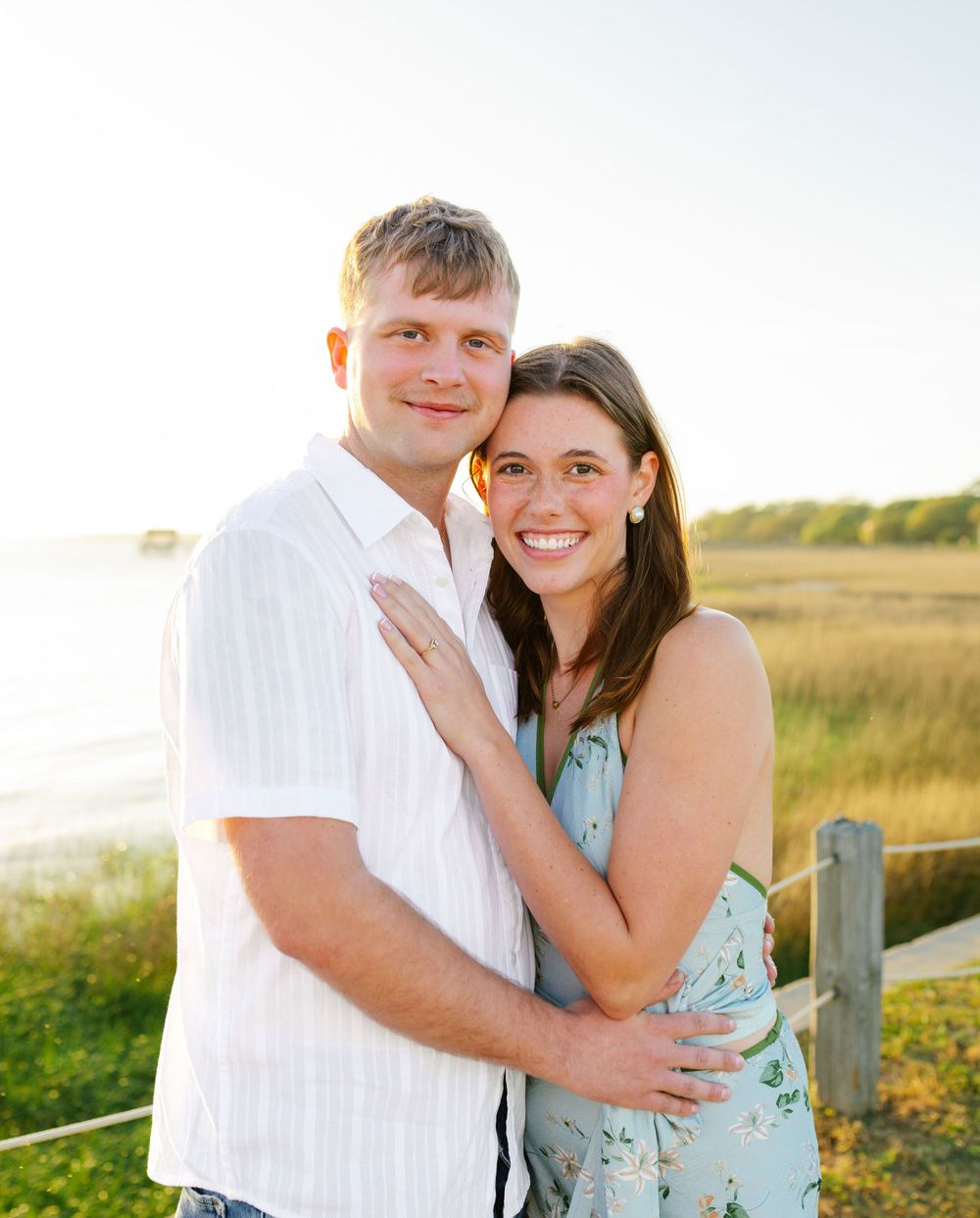 Smiling couple embraces at golden hour near a coastal marsh with a wooden fence backdrop.