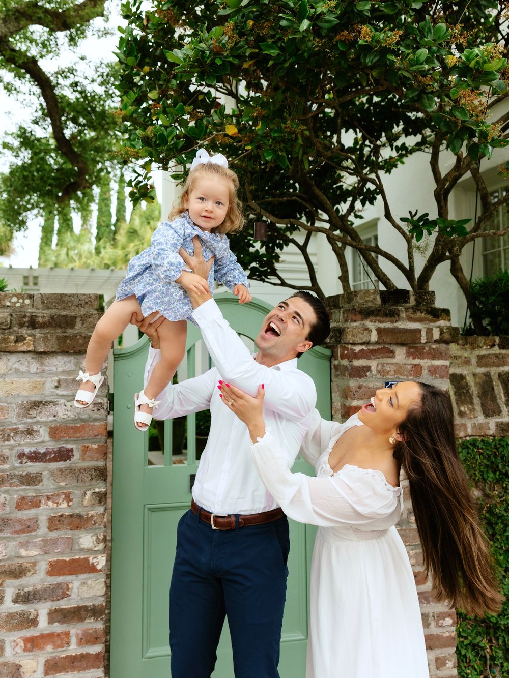 Joyful family photo with father lifting toddler girl in the air while mother watches and laughs near a green garden gate.