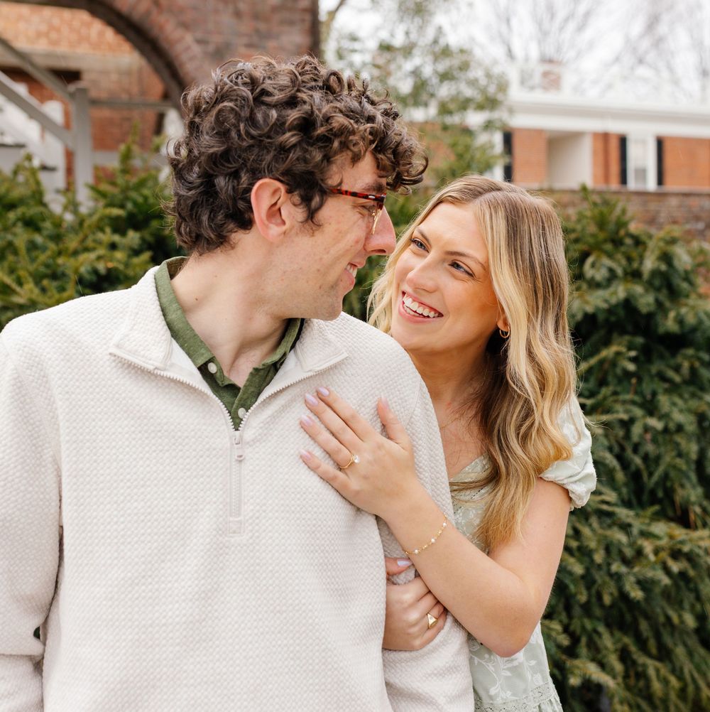A smiling couple shares a tender moment outdoors, the woman resting her hand on the man's chest near evergreen shrubs.