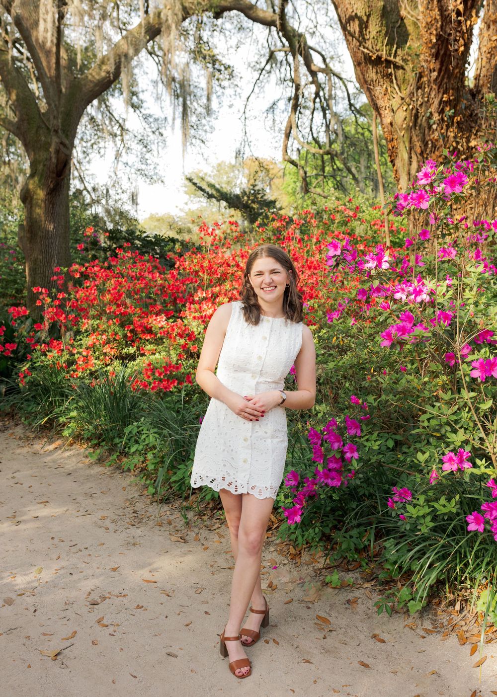 Young woman in white lace dress poses among vibrant red and pink azalea blooms on a sandy garden path.