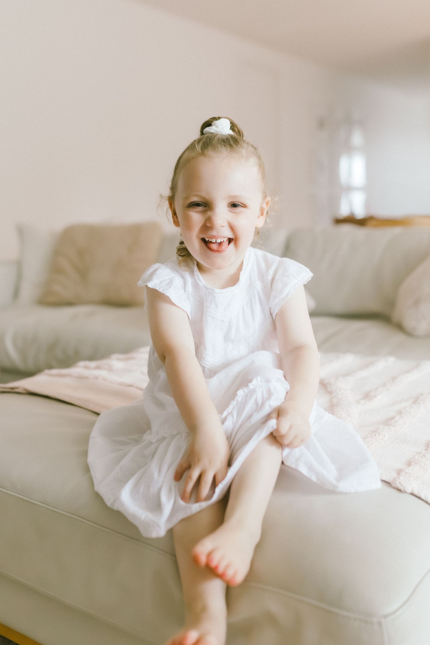 Happy young girl in white dress sitting on beige sofa, laughing joyfully with hair in bun.