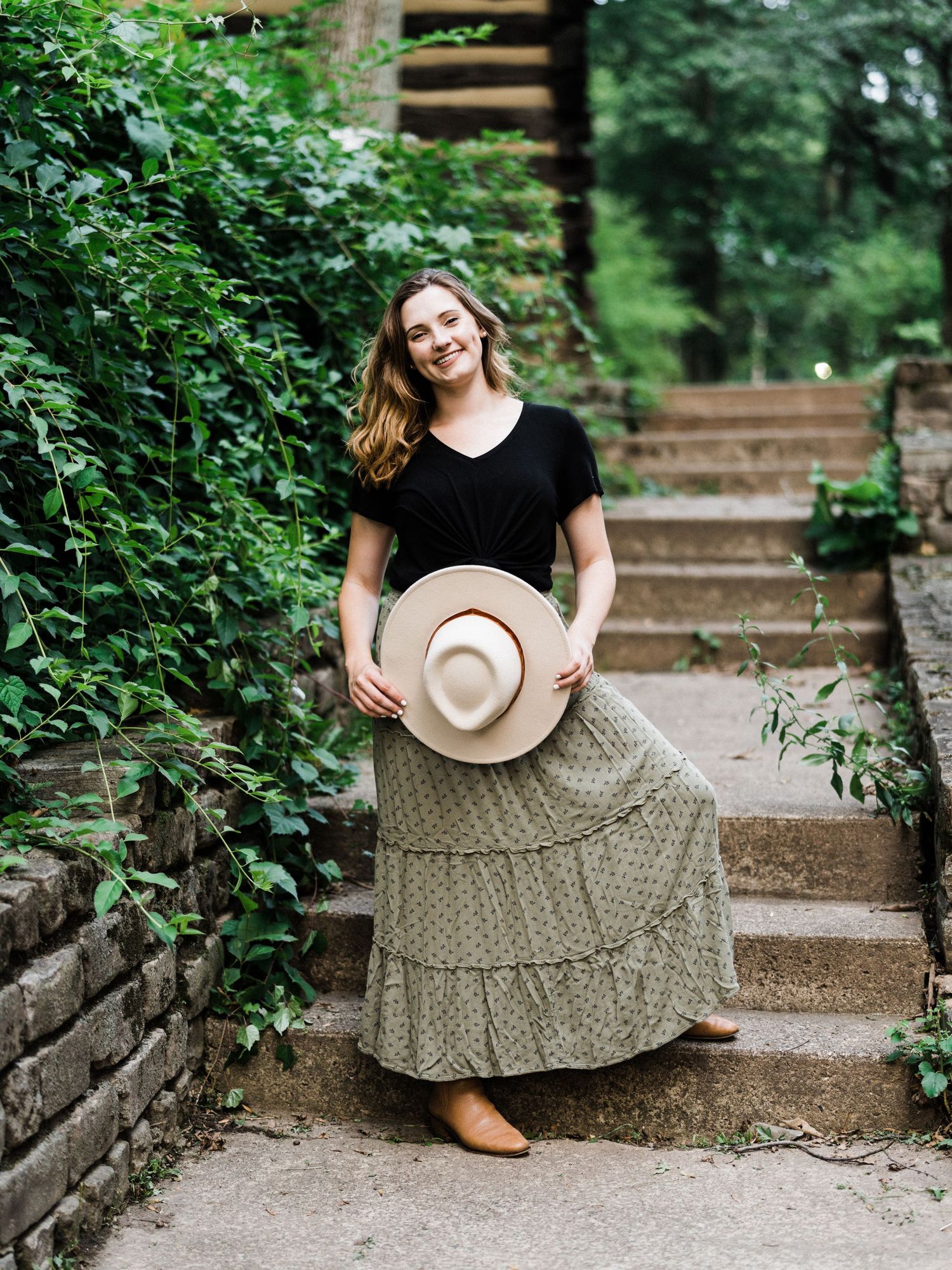Woman in black top and floral maxi skirt holding a wide-brim hat on stone steps surrounded by lush greenery.