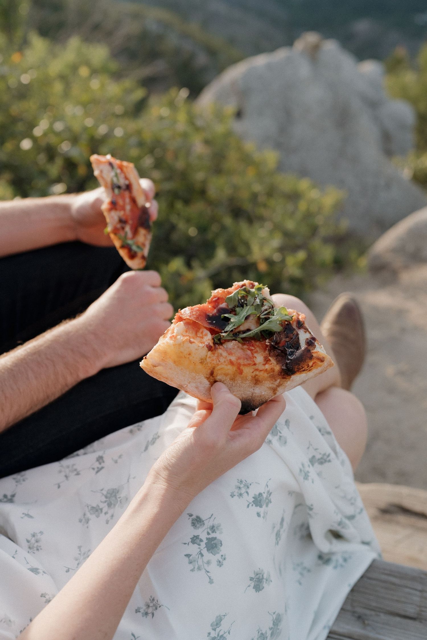 Hands holding slices of homemade pizza with melted cheese and toppings while sitting outdoors on a sunny day.