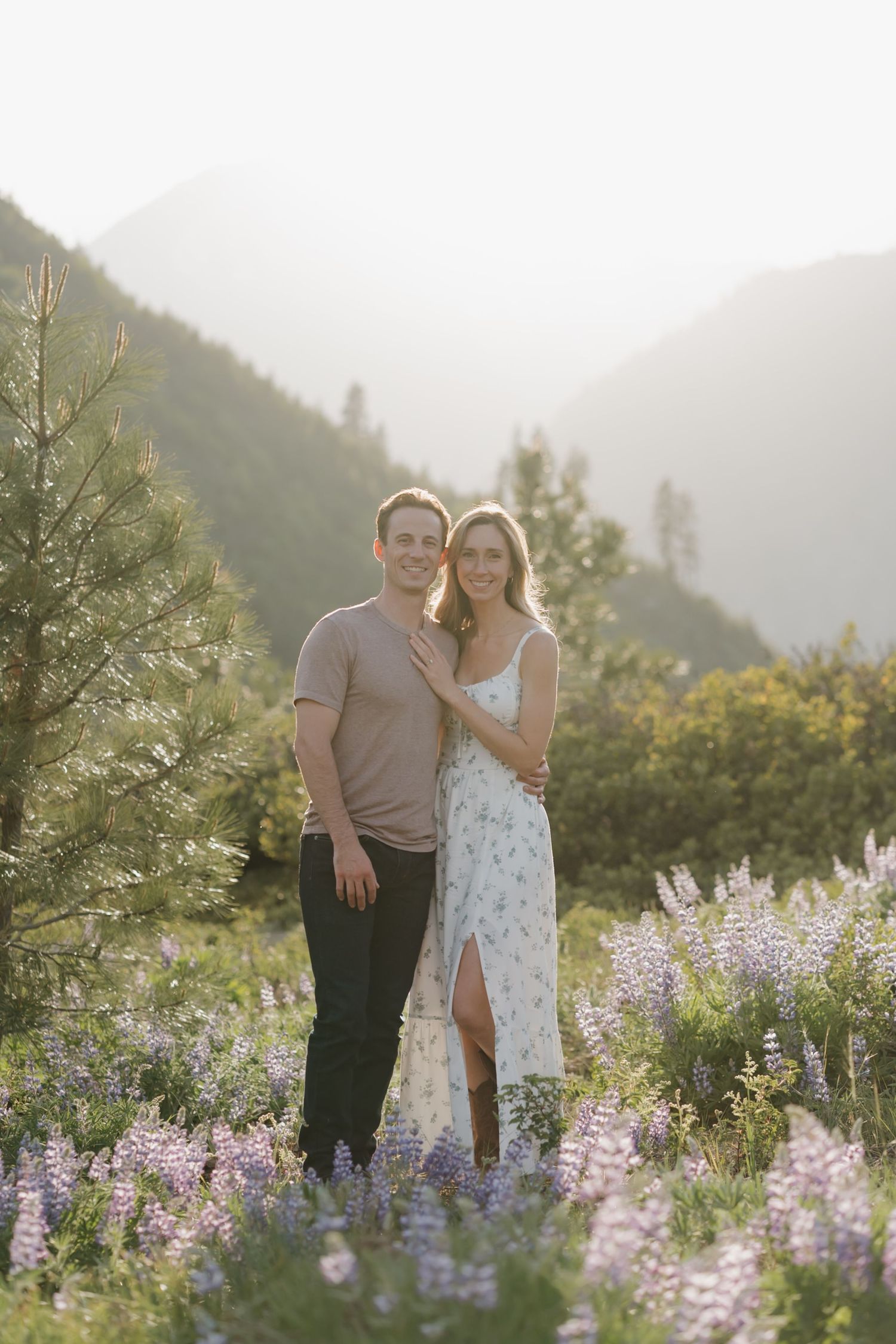 A couple stands together in a mountain meadow filled with blooming purple lupine wildflowers at sunset.