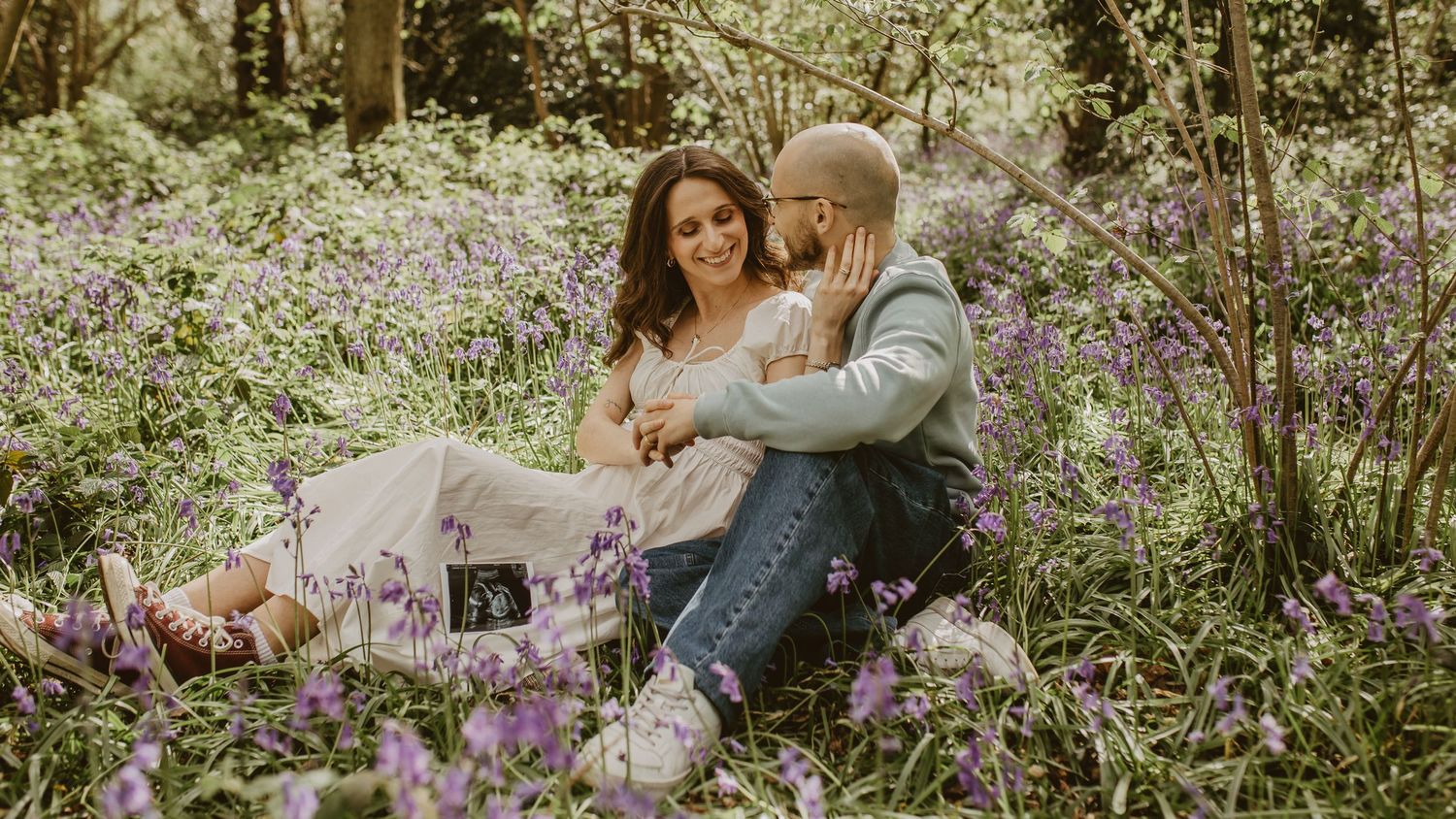 Couple sitting in a bluebell forest holding ultrasound scans, sharing a tender moment surrounded by purple wildflowers.
