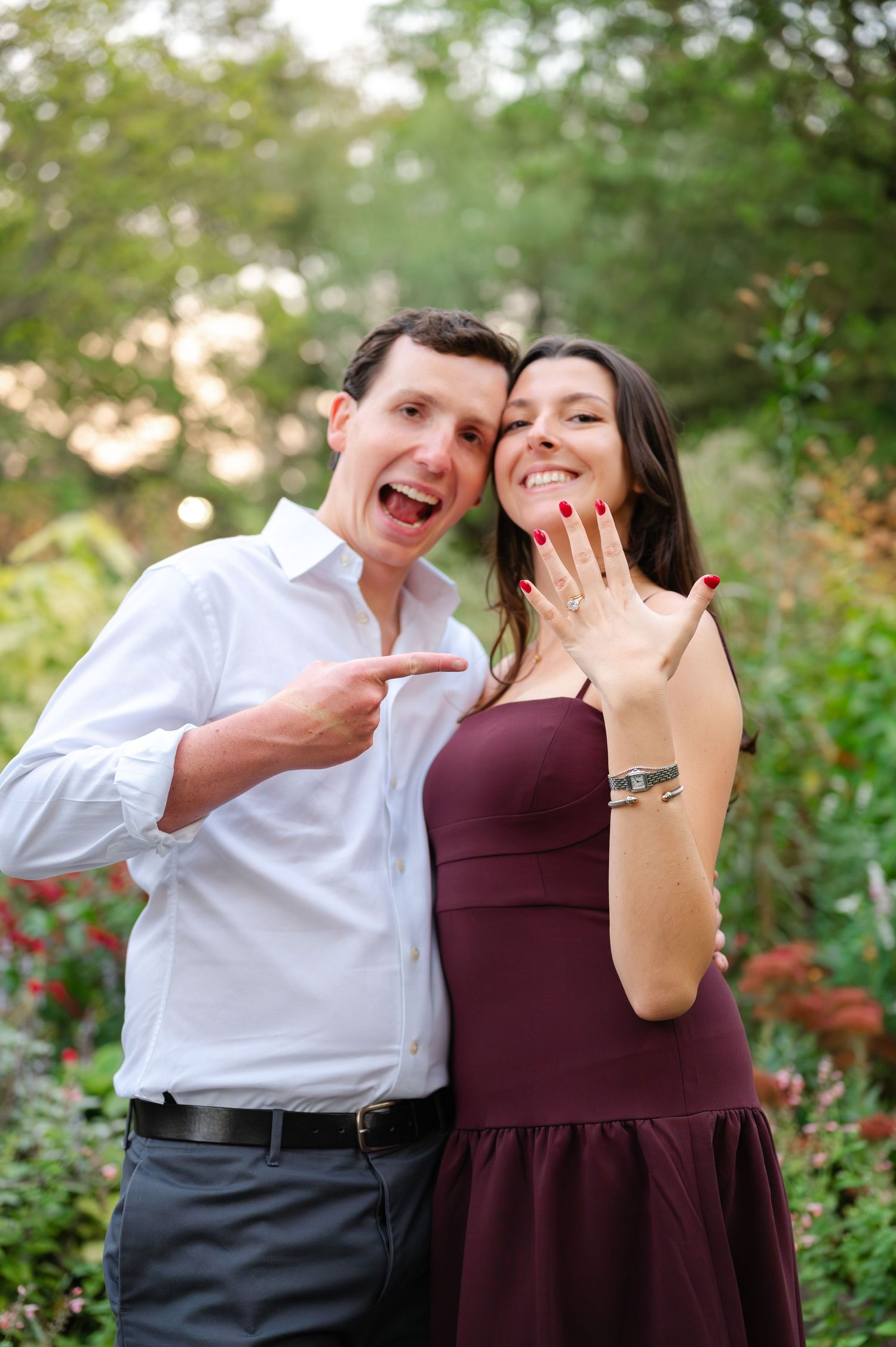 Excited couple showing off engagement ring in the Shakespeare Garden of Central Park. Man points at ring while woman displays it to camera.
