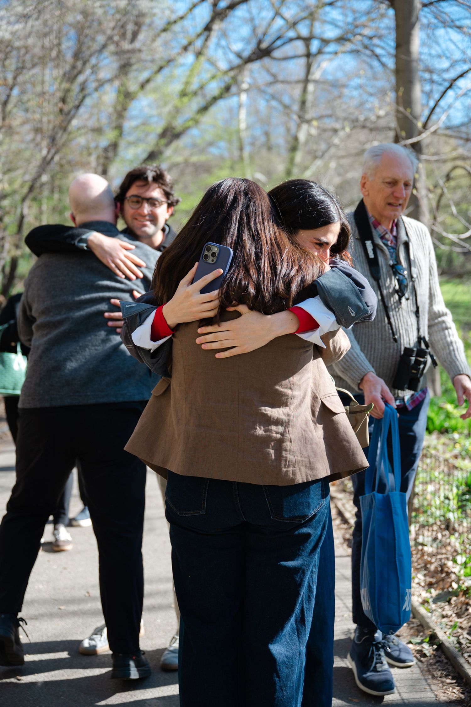 People hugging outdoors in a park during spring, with bare trees and an older man with a camera in the background.