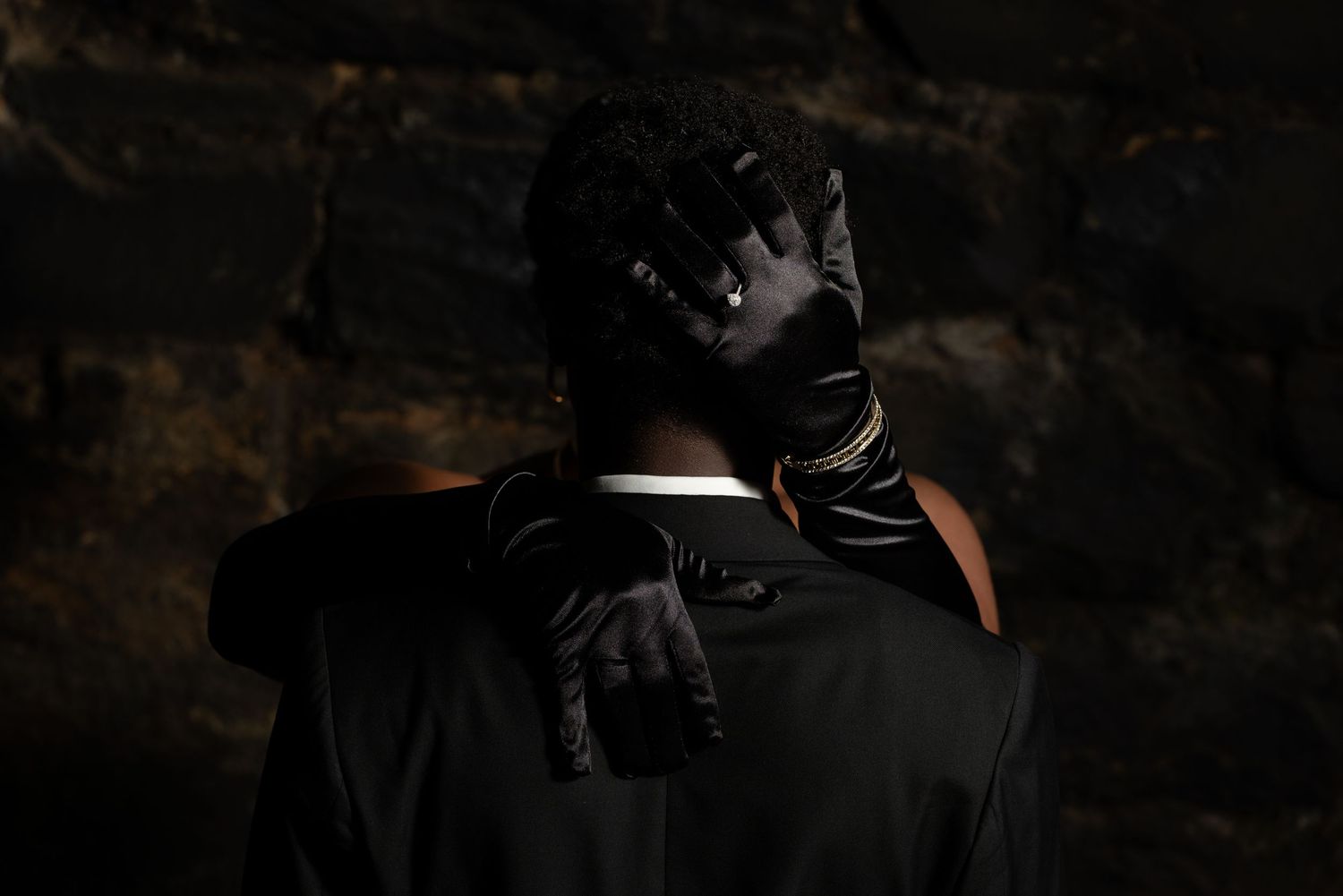 Person in black suit and leather gloves seen from behind against dark stone wall.