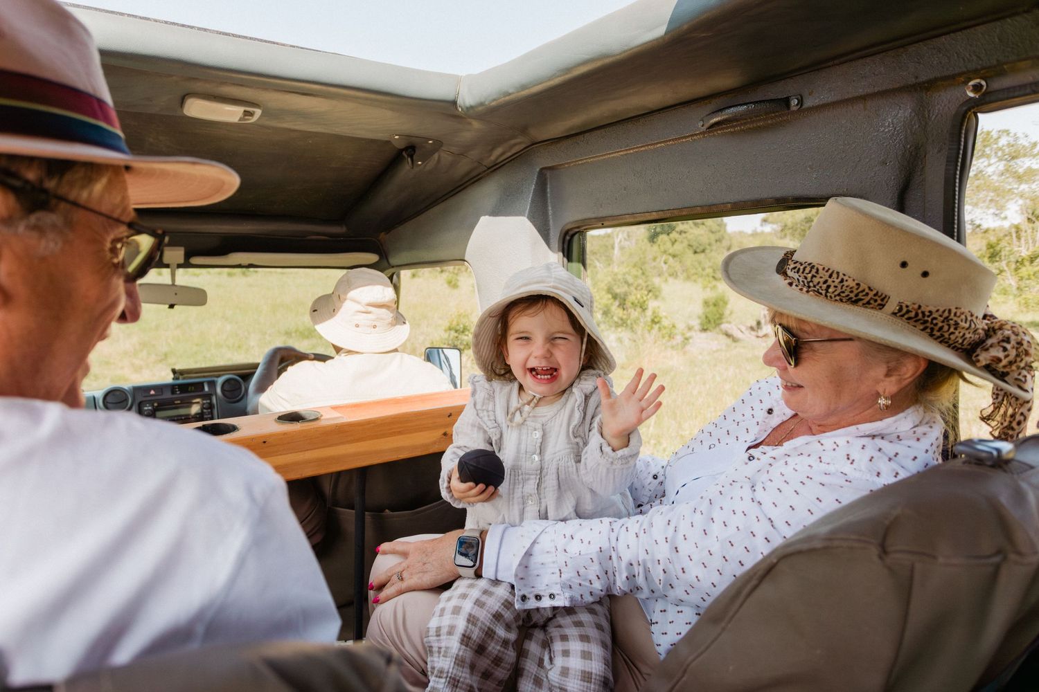 Happy baby laughing with grandparents wearing safari hats inside a safari vehicle during a family game drive.