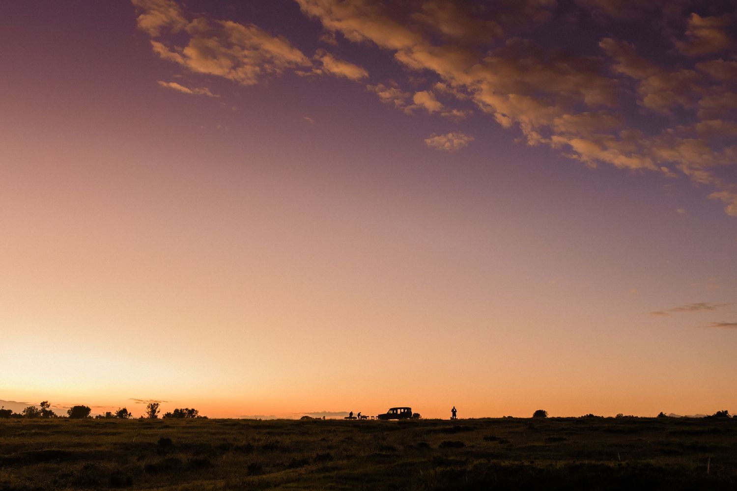 Silhouetted landscape at sunset with a vehicle and trees against a vibrant orange and purple sky.