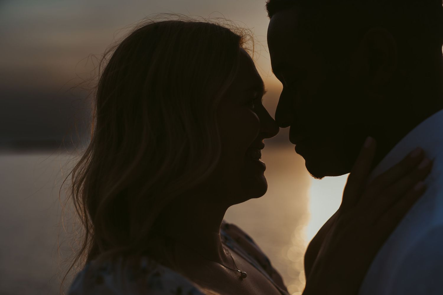 Romantic couple silhouetted against a golden sunset sky, sharing an intimate moment close together.