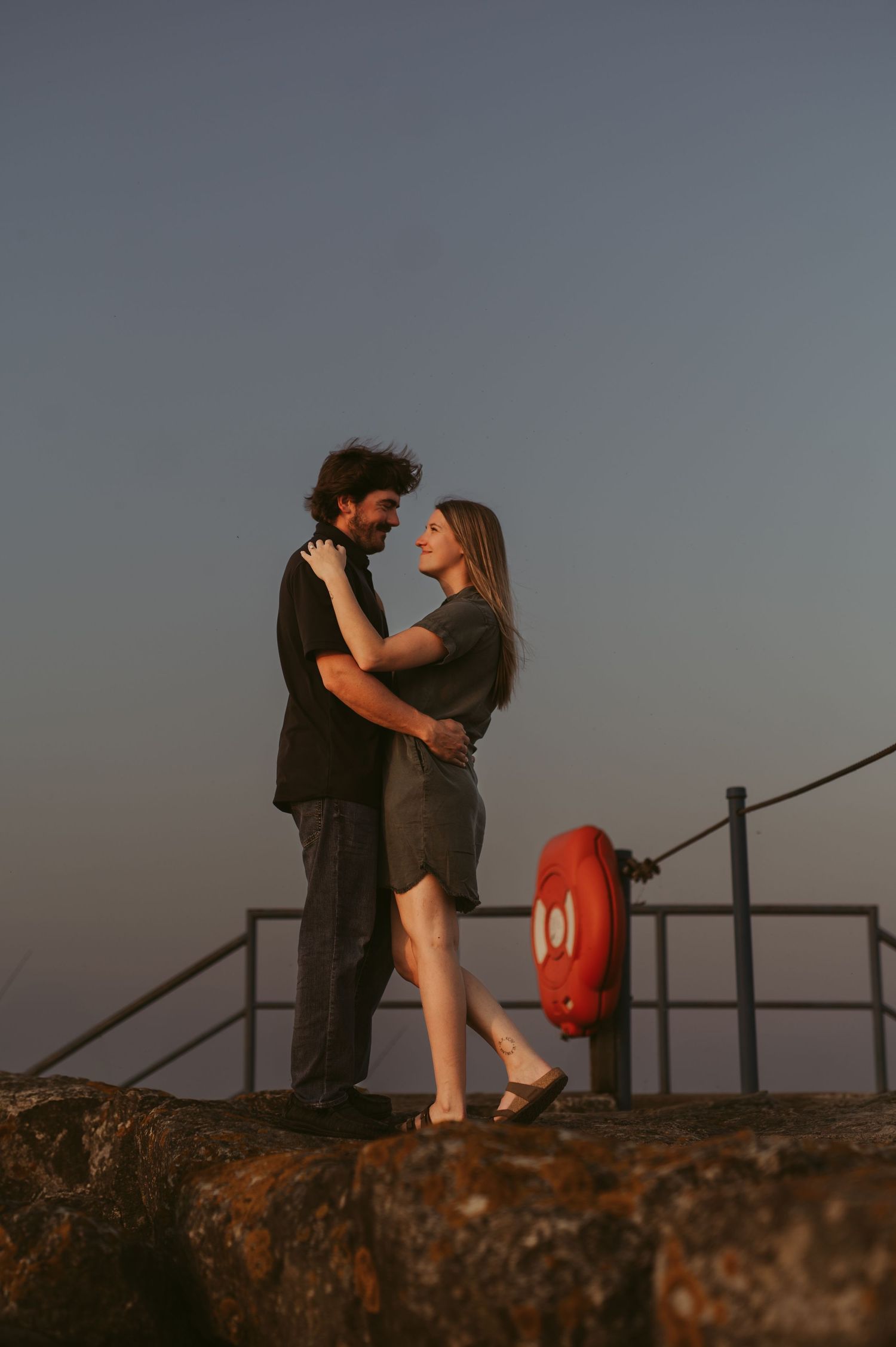 A couple embraces romantically at sunset on a rocky coastal lookout with a life preserver ring visible behind them.