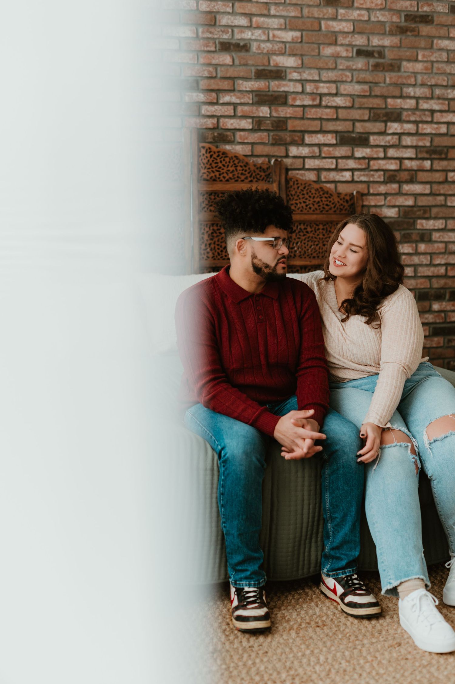 A young couple sits together, smiling and holding hands in casual jeans outfits during a couples session in Watertown Wisconsin Studio.