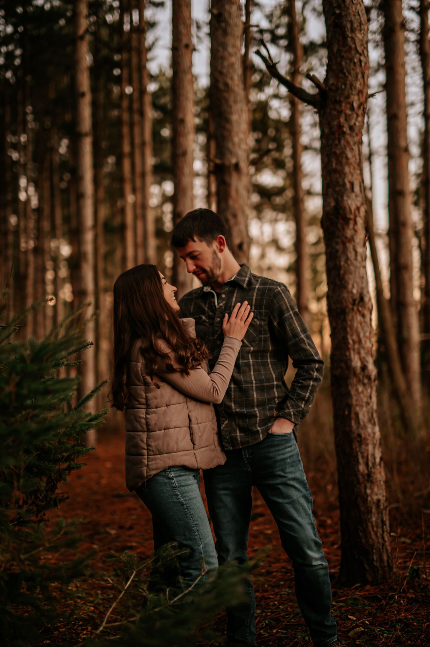 Couple sharing a romantic moment among tall pine trees in a forest during golden hour, wearing casual autumn outfits.