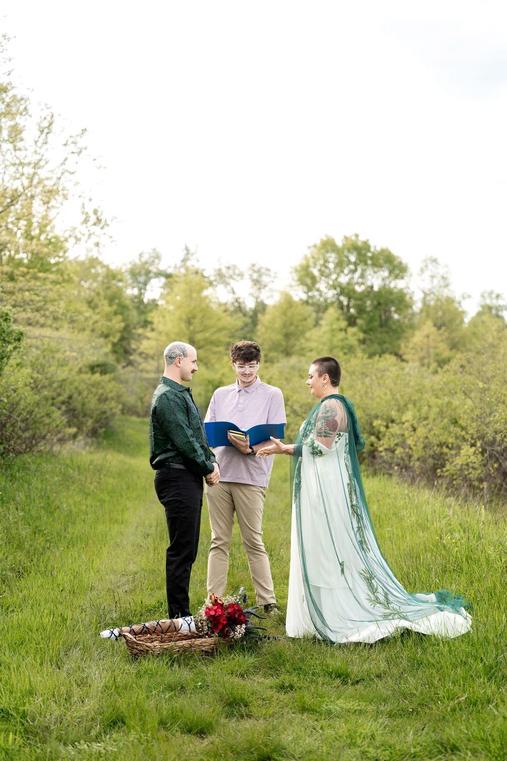 Outdoor wedding ceremony with officiant reading vows as couple stands in green field with red floral basket nearby.