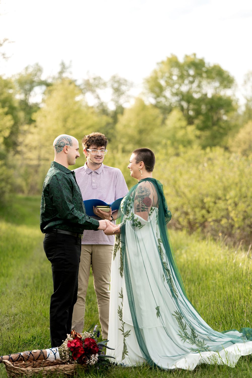 Couple exchanges vows outdoors in green field with officiant, bride wearing teal cape gown.
