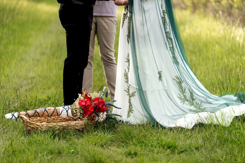 Close-up of bride's mint green embroidered gown train and red floral basket on grass at outdoor wedding.