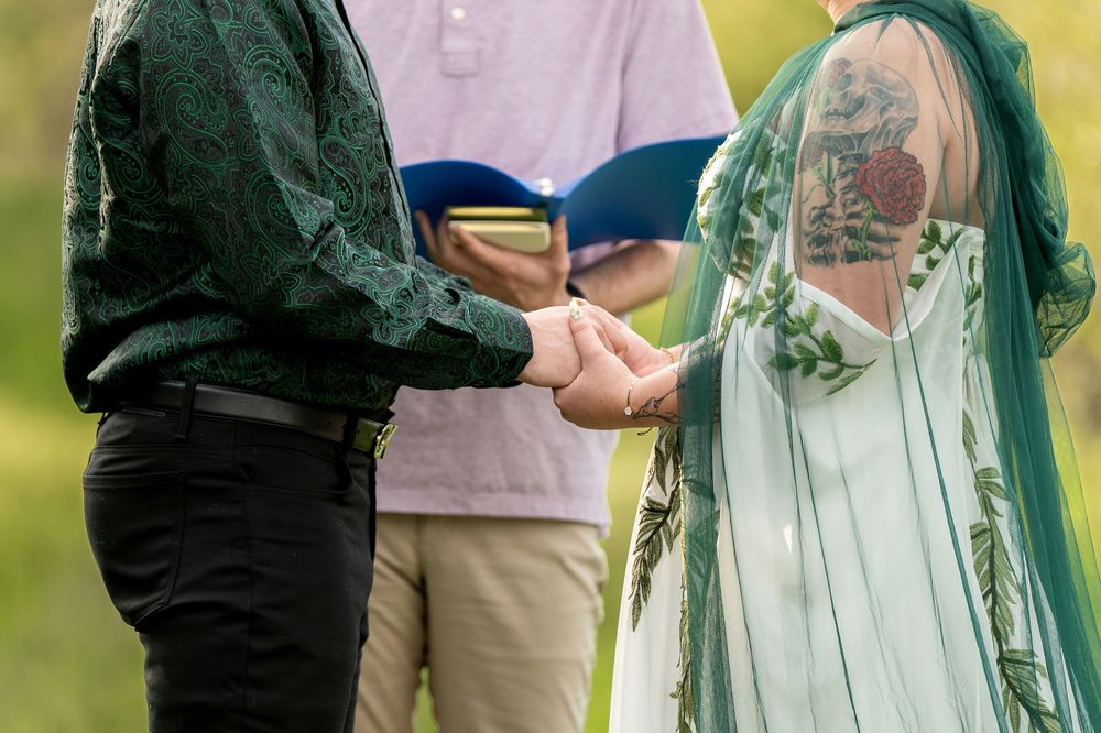 Close-up of bride and groom holding hands during outdoor wedding ceremony, officiant holding blue book in background.