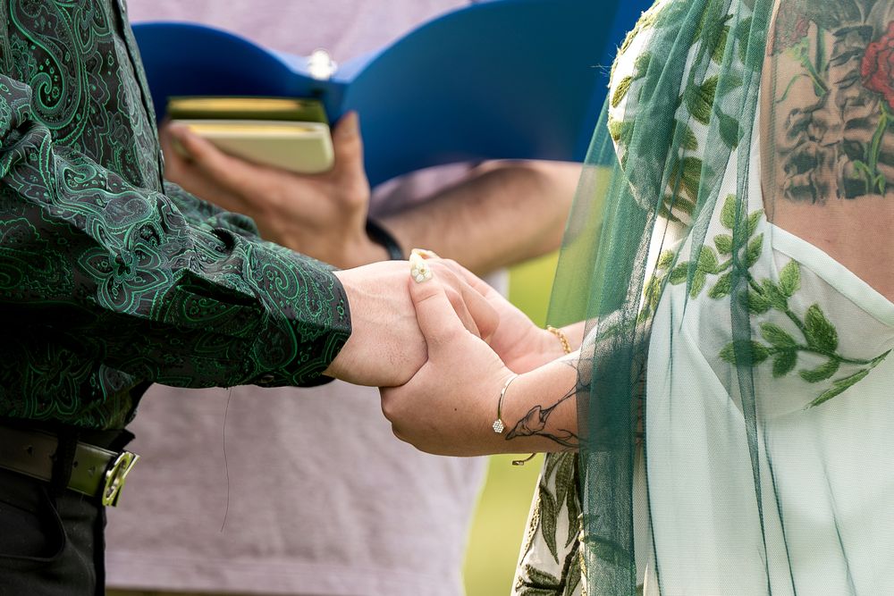 Close-up of bride and groom holding hands during outdoor wedding ceremony with green floral details.